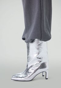 Zilveren metallic enkellaars met een spitse neus en een lage blokhak, gekenmerkt door een gekreukt textuur en een gladde afwerking. Gestyled met grijze broeken.