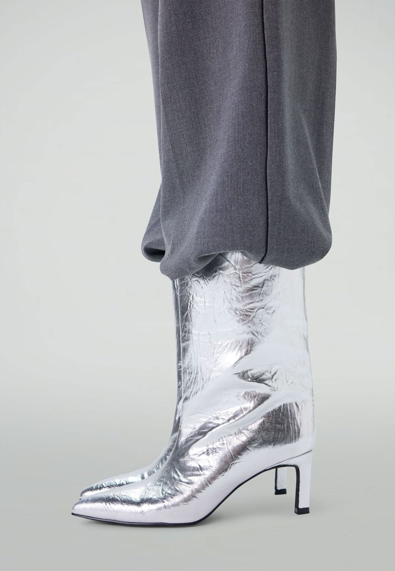 Zilveren metallic enkellaars met een spitse neus en een lage blokhak, gekenmerkt door een gekreukt textuur en een gladde afwerking. Gestyled met grijze broeken.