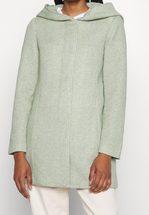 Kobieta w jasnorozzielonym wełnianym płaszczu z kapturem i przednim szwem, ubrana w białe spodnie na jednolitym tle.