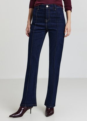 Dunkelblau gewaschene Jeans mit hoher Taille, die auffällige Vordernaht und kontrastierende gelbe Nähte aufweisen, kombiniert mit spitzen, burgunderfarbenen Absatzschuhen.