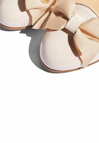 Zapatos de charol rosa pálido con un gran lazo beige texturizado, que presentan una punta redondeada y una superficie lisa y brillante.