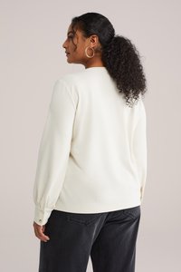 Haut à manches longues texturé ivoire avec poignets froncés et accents de boutons, associé à un pantalon gris foncé. Vue de dos.