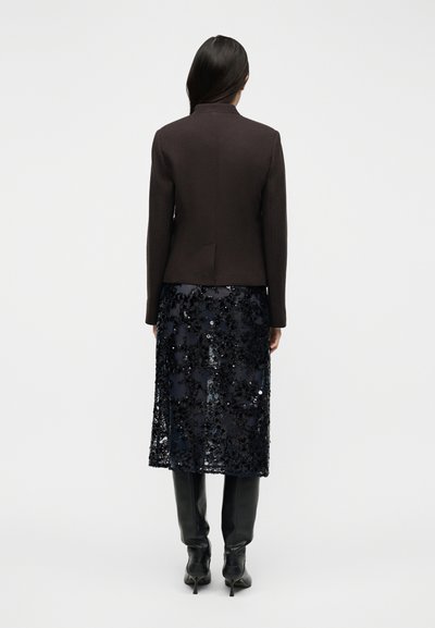 Γυναίκα που στέκεται γυρισμένη προς τα πίσω, φορώντας σκουρόχρωμο εφαρμοστό σακάκι, μια midi φούστα με παγιέτες σε ναυτικό μπλε χρώμα και μαύρες μποτάκια με τακούνι που φτάνουν μέχρι το γόνατο.