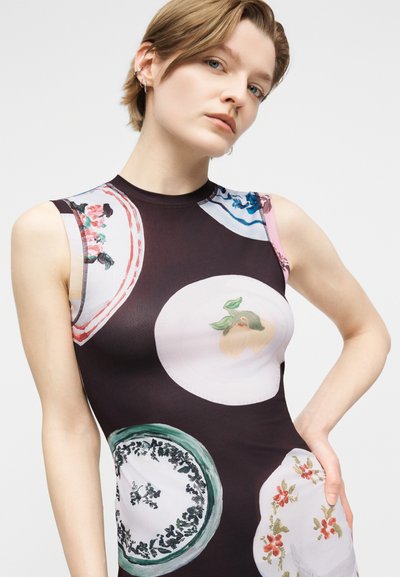 Νεαρό άτομο με κοντά μαλλιά που φοράει μαύρο αμάνικο φόρεμα με μεγάλα, πολύχρωμα λουλουδένια και κυκλικά σχέδια, ποζάροντας με το χέρι στη μέση.