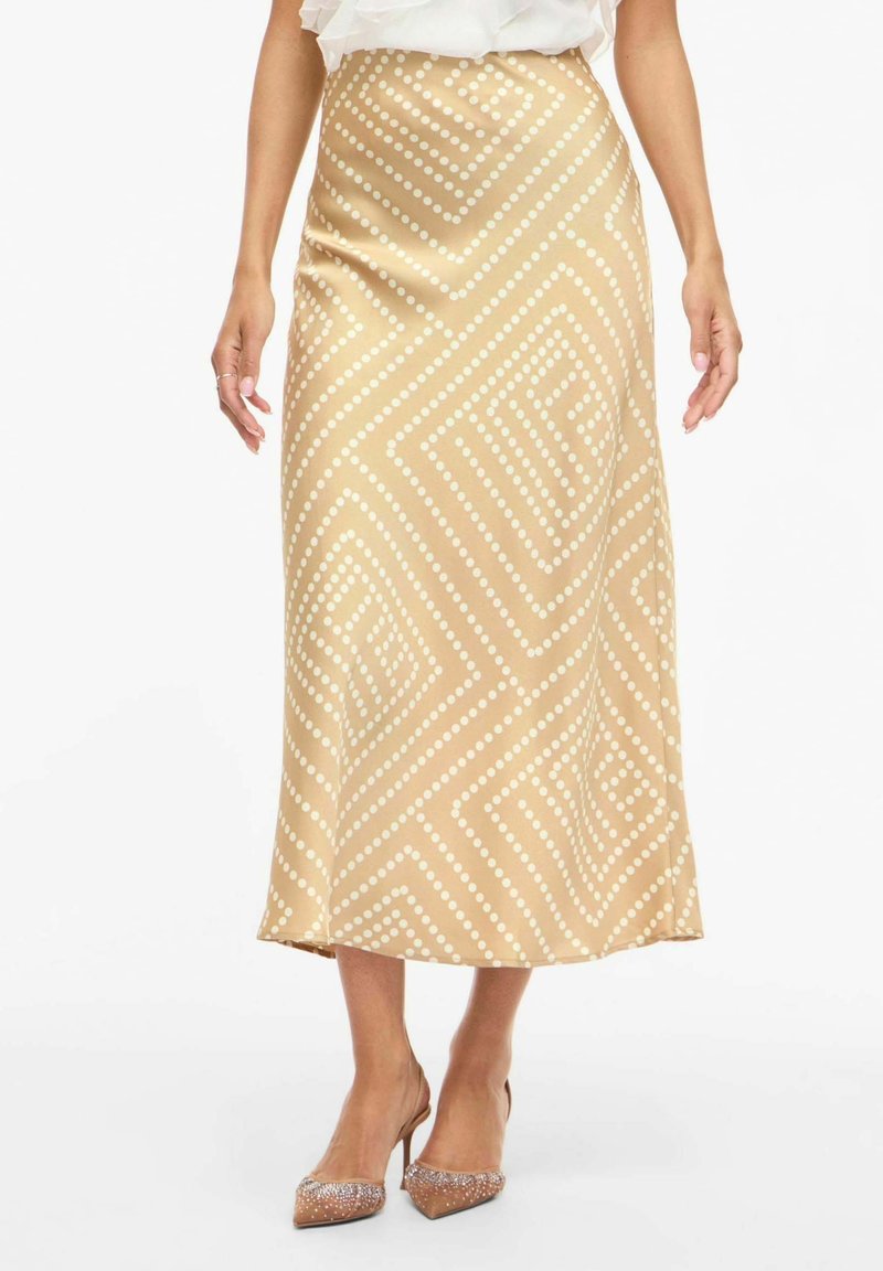 Jupe midi beige avec motifs géométriques blancs à pois, portée avec des chaussures à talons hauts transparents et un haut blanc à volants, montrée de la taille aux pieds.