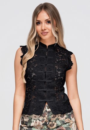 Jeune femme aux longs cheveux blonds portant un top sans manches en dentelle noire avec des boutons grenouille et un pantalon à motif floral sur un fond uni.