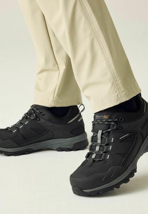 Schwarze wasserdichte Wanderschuhe mit verstärkten Sohlen und reflektierenden Schnürsenkeln, getragen mit beigen Hosen und schwarzen Socken auf hellem Hintergrund.