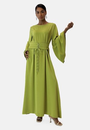 Lange, vloeiende groene jurk van lichtgewicht materiaal, met brede mouwen en een riem in de taille met een vetersluiting. Model poseert met één hand omhoog.
