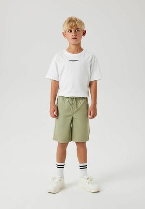 Witte katoenen t-shirt met de tekst "Studio editie", olijfgroene shorts, witte sneakers en zwartgestreepte sokken. Eenvoudige, casual outfit.