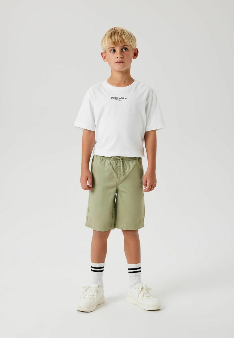 T-shirt en coton blanc avec le texte "Édition studio", shorts verts olive, baskets blanches et chaussettes rayées noires. Tenue simple et décontractée.