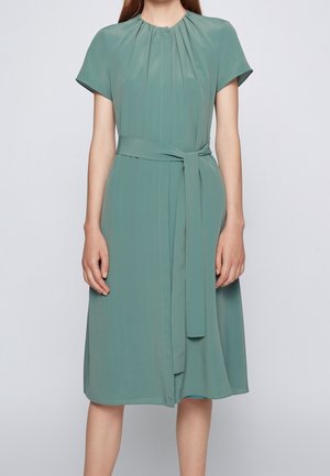 Femme portant une robe vert sauge mi-longue avec des manches courtes, un col froncé et une ceinture en tissu assortie nouée à la taille.