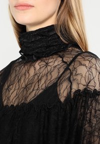 Kvinde iført sort gennemsigtig blonde bluse med høj hals, blomstermønster og flæsedetaljer, der viser overkrop og hals.