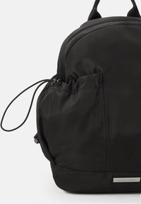 Mochila negra hecha de nylon suave, con forma redondeada, un bolsillo frontal con cordón y una etiqueta de acento plateado.
