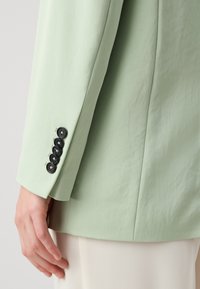 Blazer ajustado de color verde claro con una textura suave, que presenta una abertura trasera y cinco botones negros en el puño para un ajuste regulable.