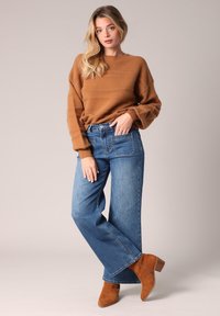 Femme aux longs cheveux blonds portant un pull marron, un jean bleu à jambes larges avec des poches avant plaquées, et des bottines en suede marron.