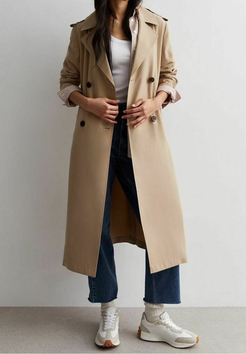 Kaki trenčkot s dvojitým zapínáním na knoflíky, velkými knoflíky, otevřeným předním dílem a proužkovanou vnitřní podšívkou. Nošený přes modré džíny a tenisky.