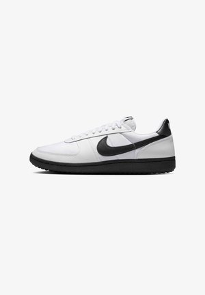 Sneaker branco com detalhes em preto, apresentando um cabedal em couro liso e uma sola em borracha texturizada. Os atacadores são brancos; o logo está em destaque.