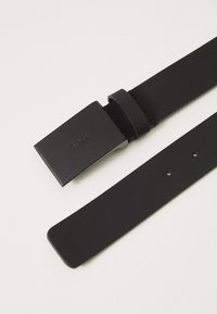 Čierny kožený opasok s obdĺžnikovou matnou prackou s embosovanou značkou, piatimi rovnomerne rozmiestnenými otvormi a čistými okrajmi.