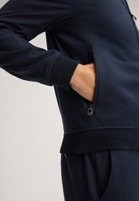 Marineblauwe zip-up jack, van opzij getoond, met een zijzak met een zilveren rits, ribgebreide manchetten en een aansluitende tailleband.
