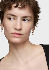 Złote kolczyki w kształcie obrączek z zawieszką, ozdobione przezroczystą koraliką i geometrycznym akcentem. Modelka ma krótkie, kręcone włosy i nosi czarną bluzkę.