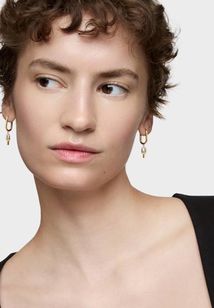 Goldene Creolen mit einem Tropfen-Design, ausgestattet mit einer klaren Perle und einem geometrischen Akzent. Das Model hat kurze, lockige Haare und trägt ein schwarzes Oberteil.