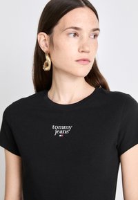 Μαύρο βαμβακερό μπλουζάκι με κοντά μανίκια, με λευκό λογότυπο "tommy jeans" και μία μικρή χρωματιστή λεπτομέρεια. Μοντέλο που φορά αχάριστα σκουλαρίκια σε χρυσό τόνο.