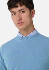 Pastelově modrý pletený svetr s kulatým výstřihem, s texturovaným, měkkým povrchem. Pod ním je světle modrá pruhovaná košile s límečkem.