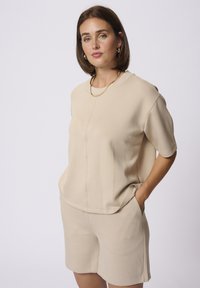 Ensemble bi-matière beige clair composé d'un haut à manches courtes au fit décontracté avec poches latérales, associé à un short assorti. Texture lisse.