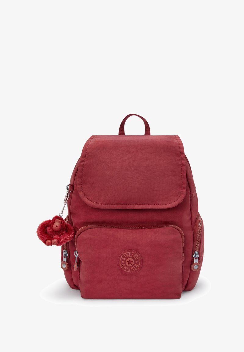Czerwony plecak z materiału z klapą, przednią kieszenią na zamek i dekoracyjnym breloczkiem małpki. Ma naszywkę z logo i szczegóły przeszycia.