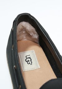 Chaussure noire à enfiler avec doublure intérieure douce, étiquette en tissu affichant "UGG" et couture détaillée. Texture extérieure lisse avec des bords arrondis.