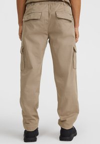 Beige cargo-housut rennolla leikkauksella, joustovyötäröllä, sivutaskuilla ja sileällä pinnalla. Yhdistetty mustiin kenkiin.