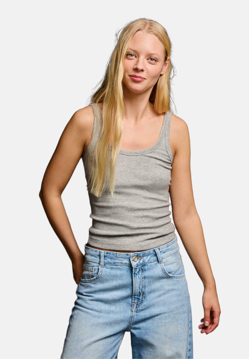 Jeune femme aux longs cheveux blonds, portant un débardeur gris et un jean bleu clair, debout, une main dans la poche, fond neutre.