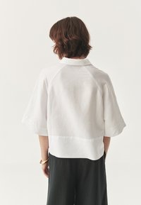 Camicetta bianca in lino a maniche corte con colletto, caratterizzata da una vestibilità comoda e un orlo posteriore leggermente più lungo. Indossata con pantaloni neri, presenta un design minimalista.