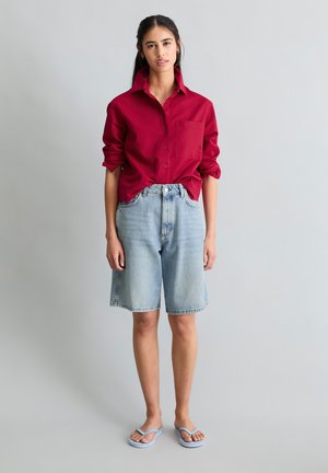 Junge Frau steht und trägt ein rotes Langarmshirt mit hochgekrempelten Ärmeln, helle Jeans-Shorts bis zum Knie und hellblaue Flip-Flops.