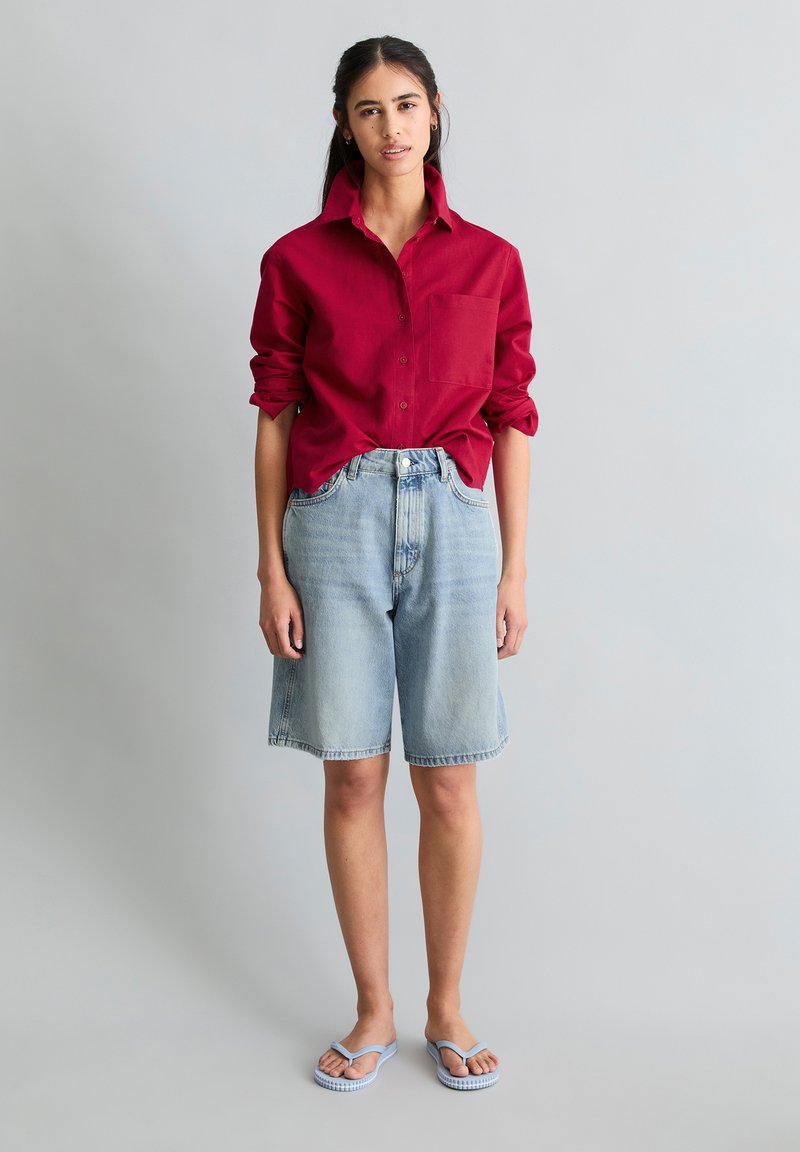 Junge Frau steht und trägt ein rotes Langarmshirt mit hochgekrempelten Ärmeln, helle Jeans-Shorts bis zum Knie und hellblaue Flip-Flops.