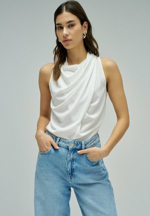 Femme aux cheveux foncés portant un haut blanc sans manches drapé et un jean bleu clair taille haute, les mains dans les poches, devant un fond gris uni.