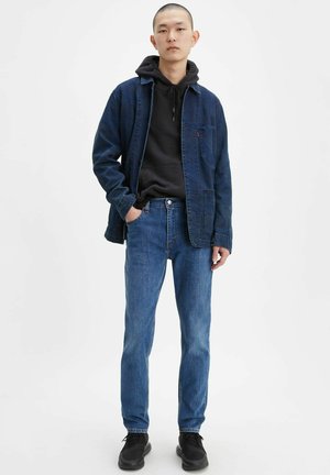 Veste en denim bleu foncé, sweat à capuche noir en dessous, jeans bleus et baskets noires ; comporte des poches et une coupe décontractée.