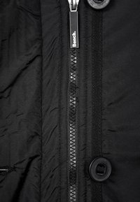 Černá zipová bunda s hladkou texturou, kovovým zipem označeným "Bench" a zaoblenými černými knoflíky na léze.