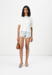 Witte cropped t-shirt met bloemenborduring, gecombineerd met versleten denim shorts. Accessoires bestaan uit een bruine handtas en grijze puntige flats.