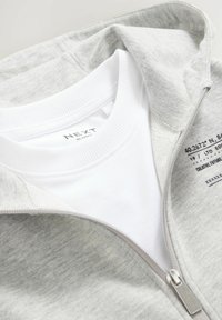 Grå zip-up hoodie lagd over en hvid t-shirt. Hoodie'en har en glat tekstur, en frontlynlås og mærkning indvendigt.