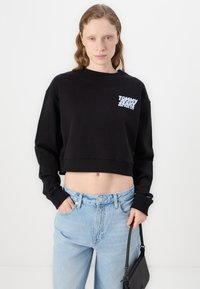Černá zkrácená mikina s žebrovanými manžetami a lemem, s logem "TOMMY HILFIGER" v modré barvě na hrudi. V kombinaci se světle modrými džíny s vysokým pasem.