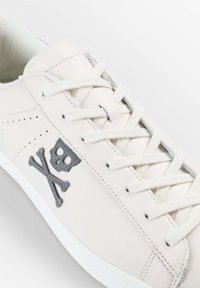 White leather sneaker featuring gray skull and crossbones embroidery, flat white laces, and perforated details on the upper.