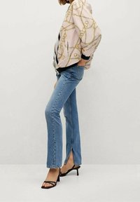 Jasnoróżowa kurtka bomber z wzorem złotego łańcucha, zestawiona z niebieskimi, rozszerzanymi dżinsami z przednim rozcięciem oraz czarnymi sandałkami na paski.