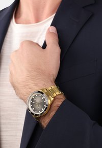 Goldene Armbanduhr mit einem runden grauen Zifferblatt, schwarzem Ring, Stundenmarkierungen und goldenem Armband, getragen an einer hellhäutigen Hand unter einer marineblauen Jacke.