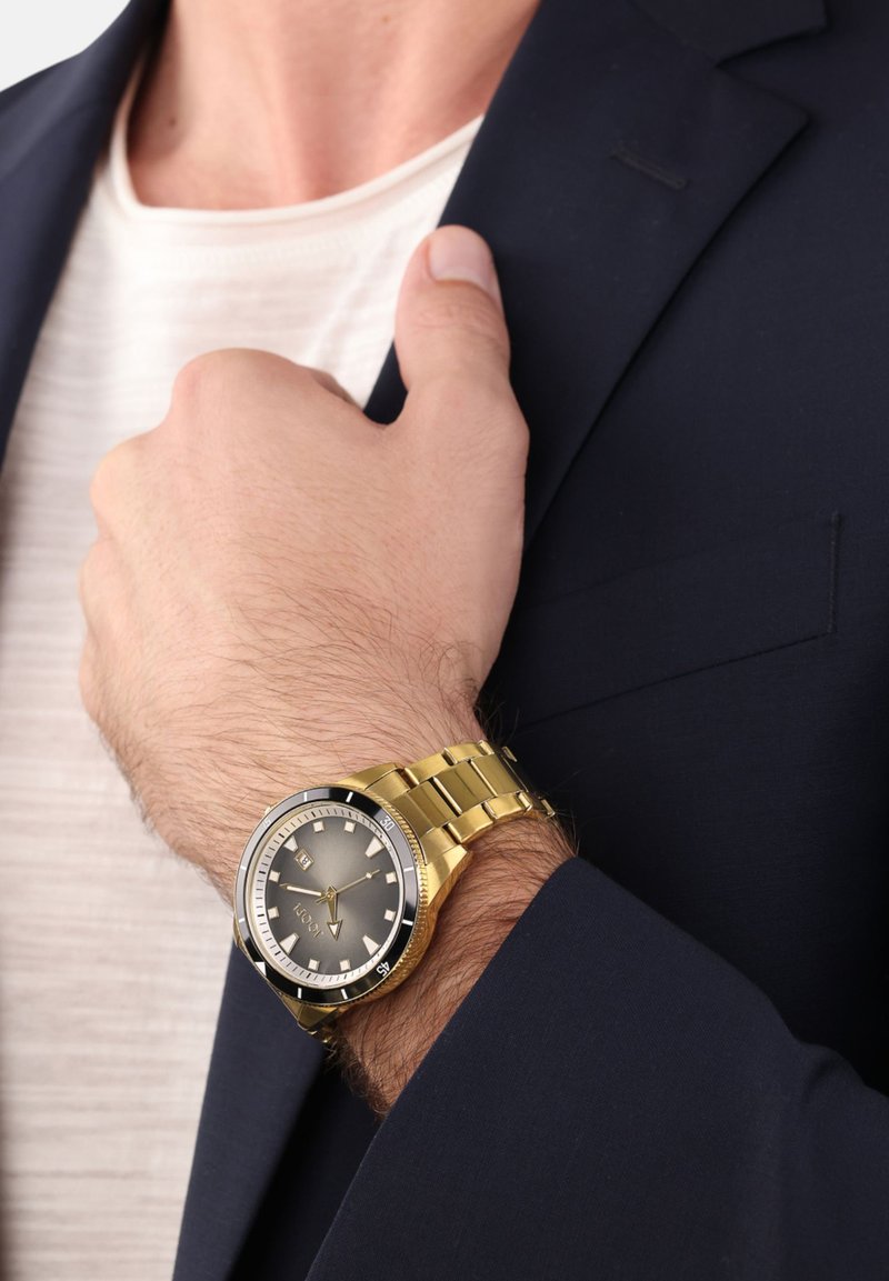 Goldene Armbanduhr mit einem runden grauen Zifferblatt, schwarzem Ring, Stundenmarkierungen und goldenem Armband, getragen an einer hellhäutigen Hand unter einer marineblauen Jacke.