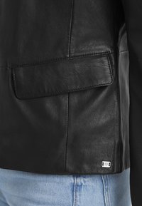 Schwarze Lederjacke mit einer aufgesetzten Tasche, glatter Textur und Nähdetails; enthält ein kleines Markenetikett am Saum.