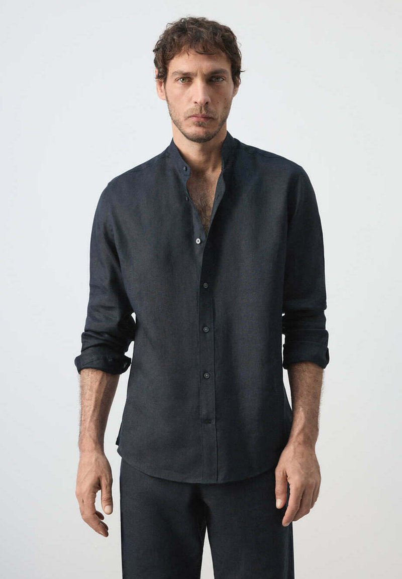 Homme aux cheveux courts et bouclés portant une chemise sombre à manches longues boutonnée avec un col mandarin et un pantalon sombre assorti, sur un fond uni.