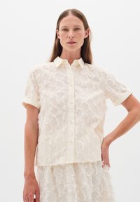 Cream-colored button-up shirt with short sleeves, featuring a textured, fringed fabric pattern and a collar.