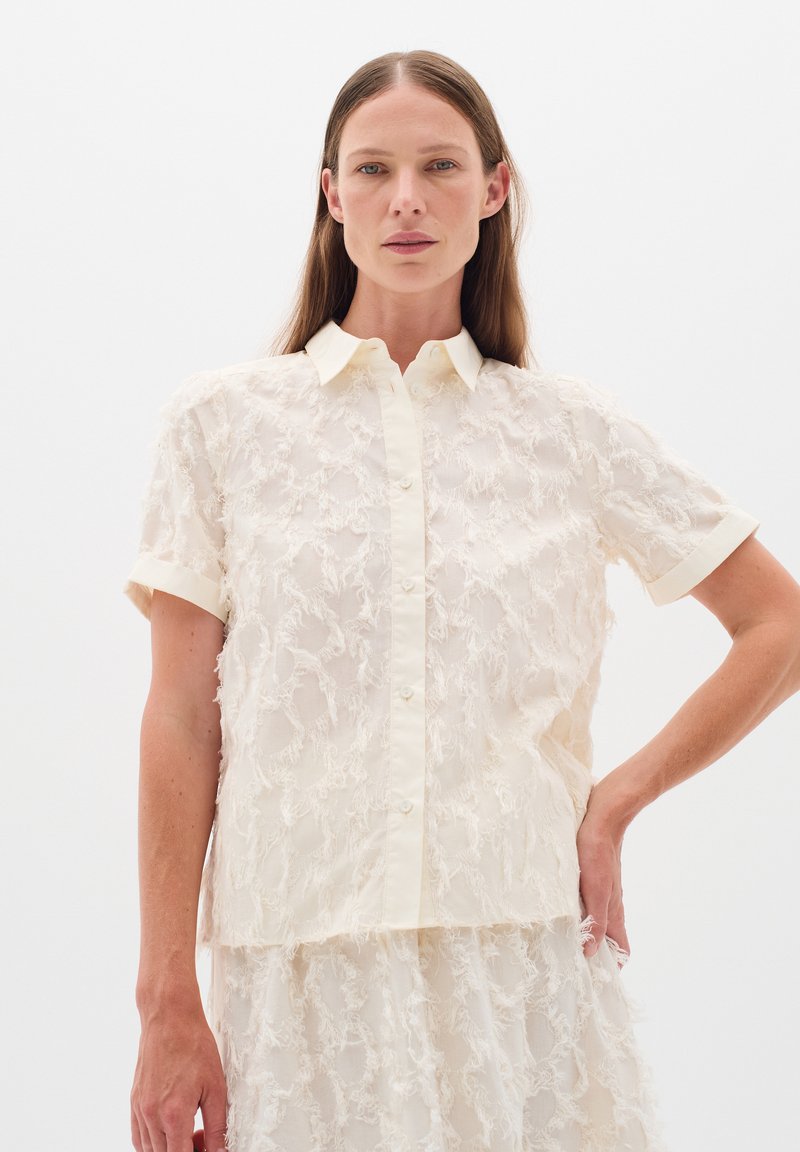Cream-colored button-up shirt with short sleeves, featuring a textured, fringed fabric pattern and a collar.