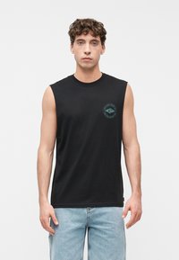 Νεαρός άνδρας με σγουρά μαλλιά που φοράει μαύρο αμάνικο πουκάμισο με ένα μικρό τιρκουάζ κυκλικό λογότυπο και ανοιχτόχρωμο τζιν.